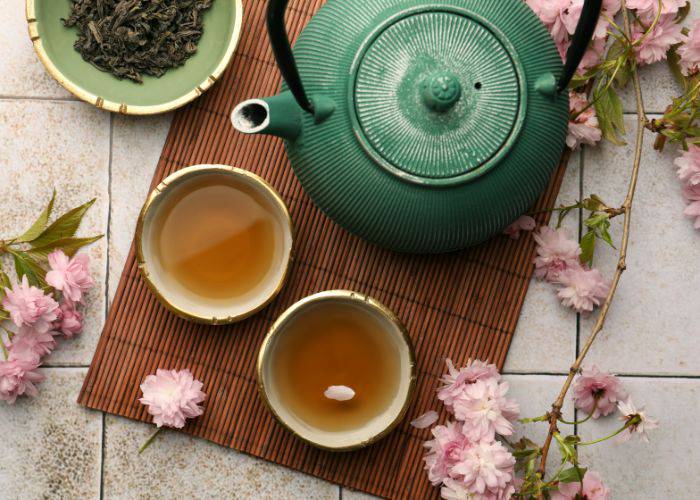 A serving of cherry blossom tea in two bowls, surrounded by a Japanese tea pot, tea leaves, and sakura petals.